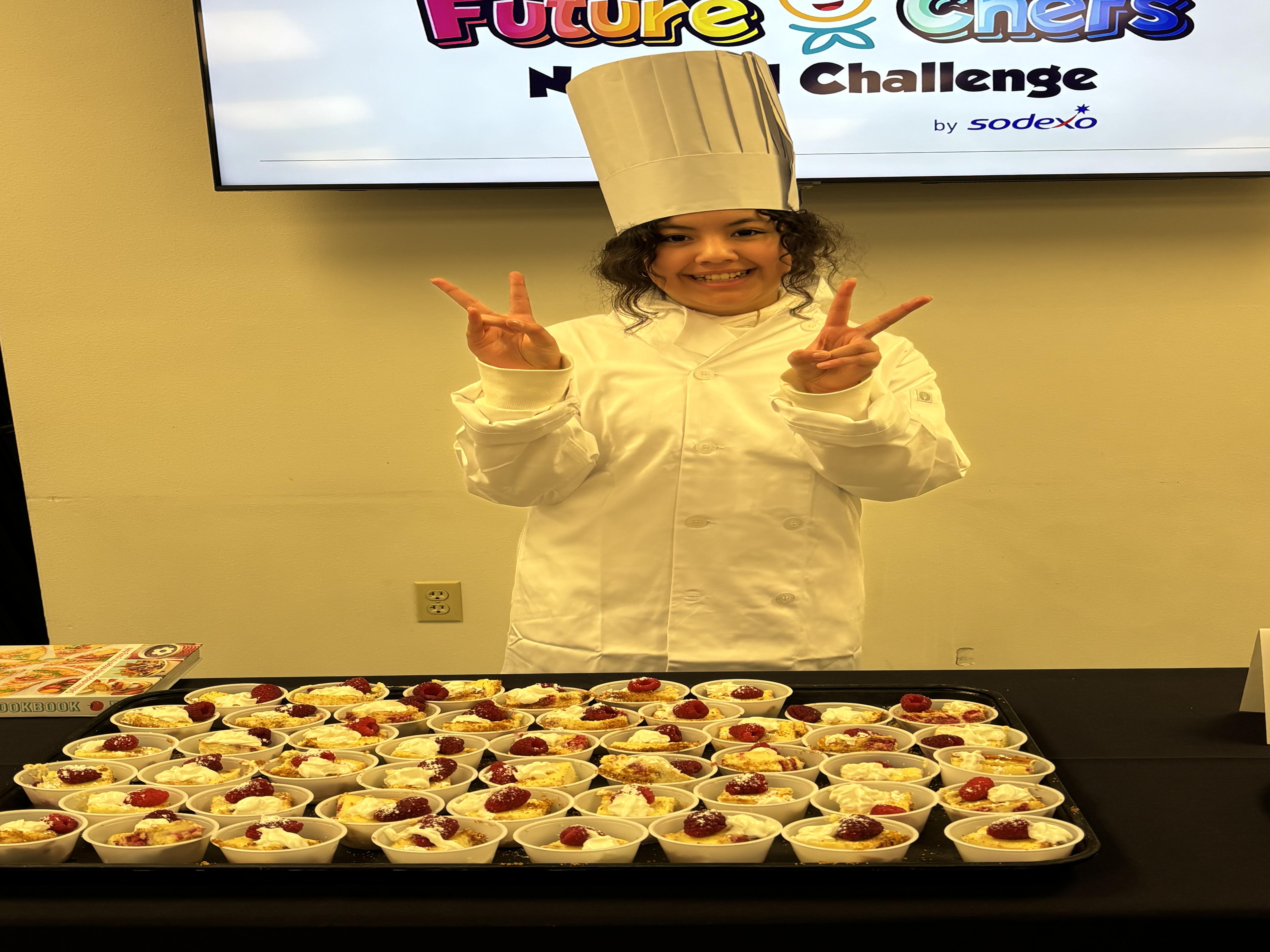 Student in a chefs outfit with a white coat and chef hat is smiling in front of a display of desserts on a black table. She is giving the peace sign with both hands.